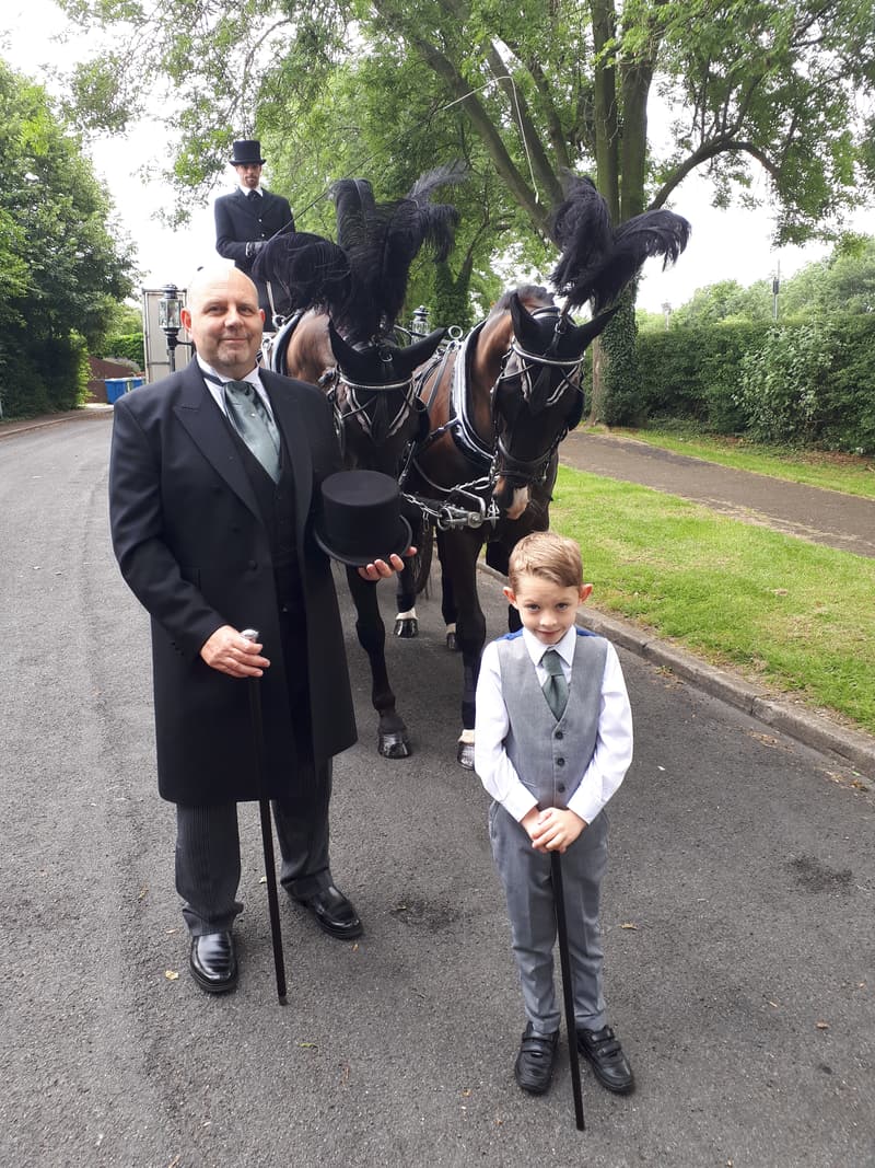 Steve Grocutt in traditional mourning dress and top hat with a young page boy, in front of a horse-drawn hearse