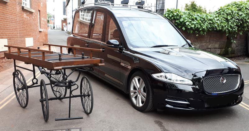 A black Jaguar hearse parked beside a traditional wooden wheeled funeral bier