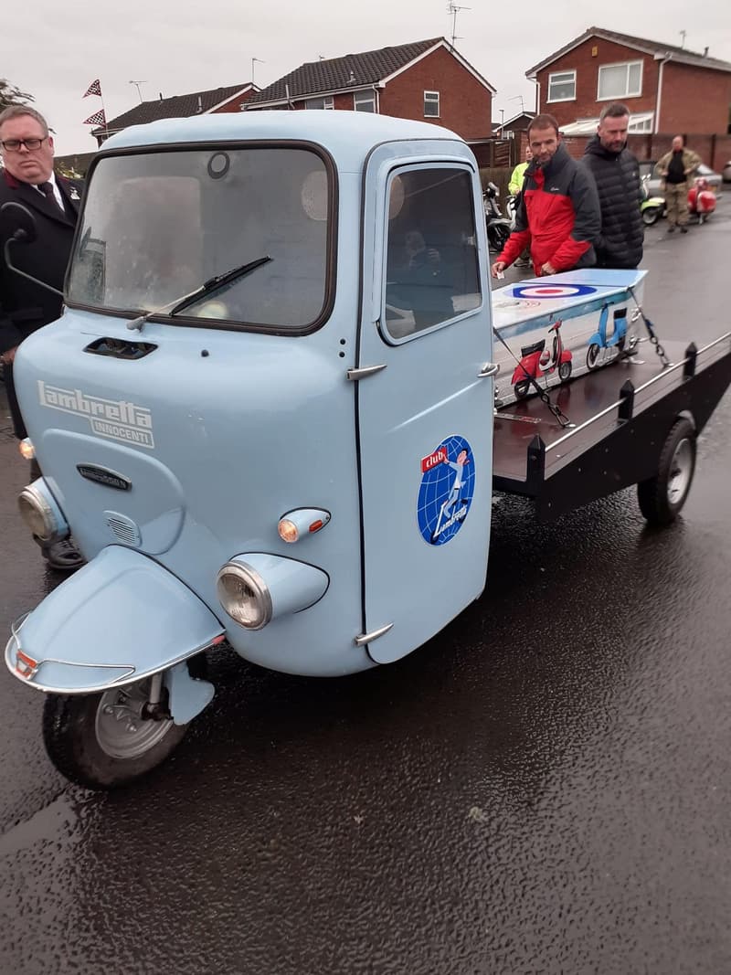 A pale blue 1967 Lambretta three-wheeler hearse carrying a coffin, with mourners alongside