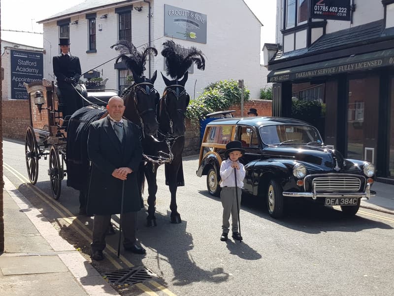 A horse-drawn hearse and a black Morris Minor hearse on a town street, with a funeral director and a page boy