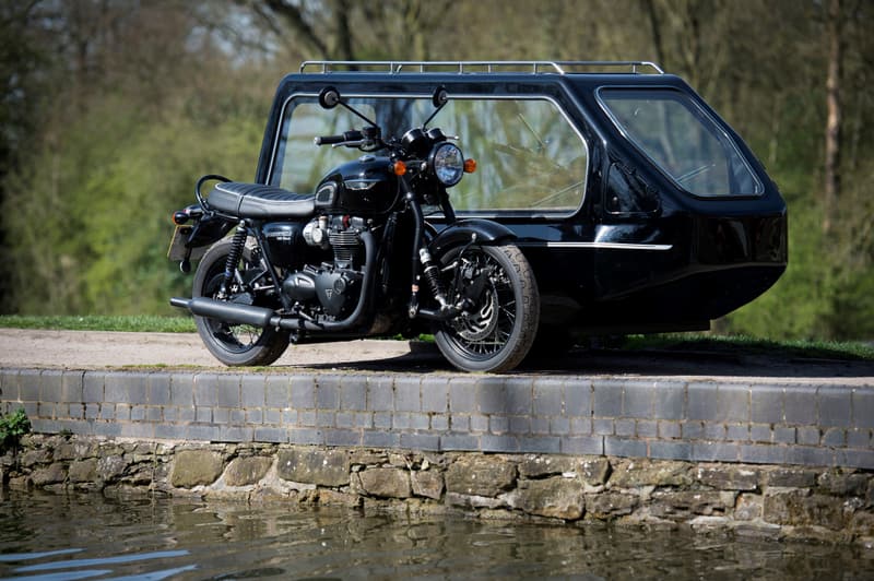 A black motorcycle hearse with a glass-sided sidecar, parked beside still water